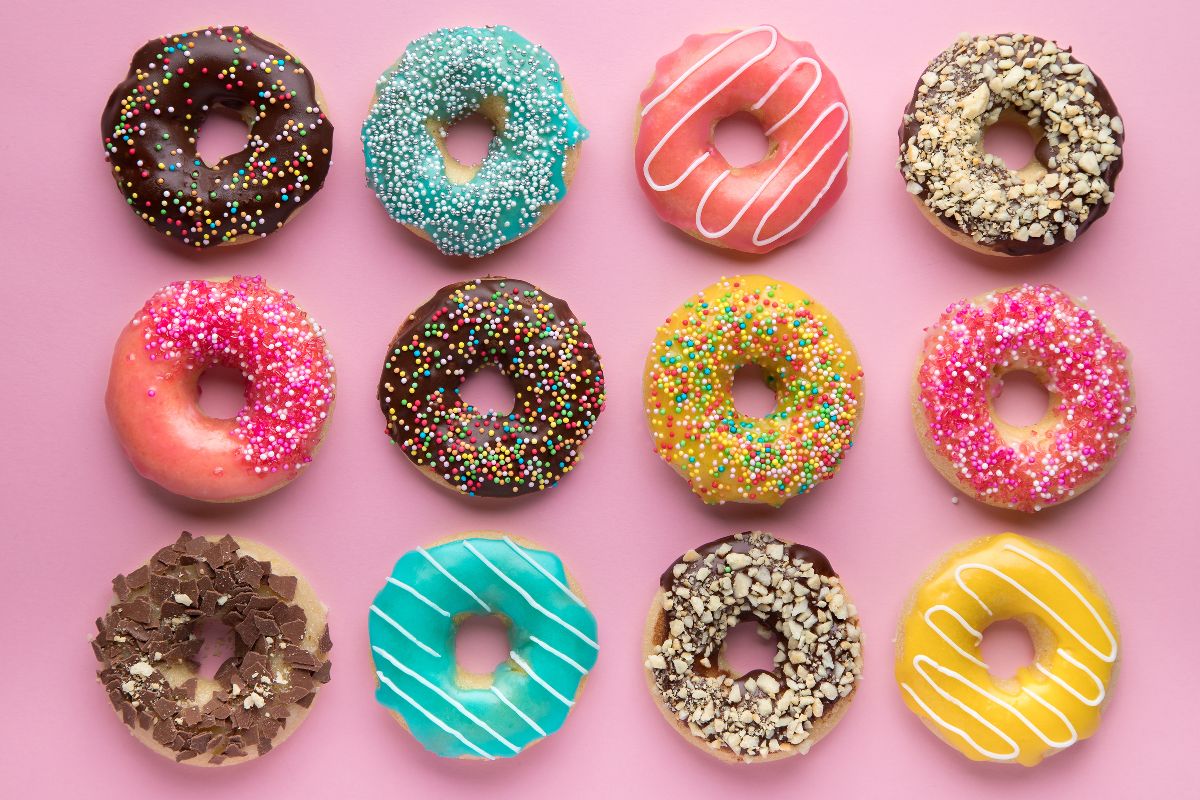 A variety of twelve different colored/flavored donuts in front of a pink background 