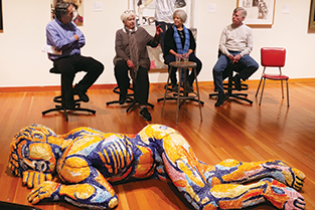 Group of four artists sit on stools talking, with large, colorful human sculpture laying on the floor in front of them