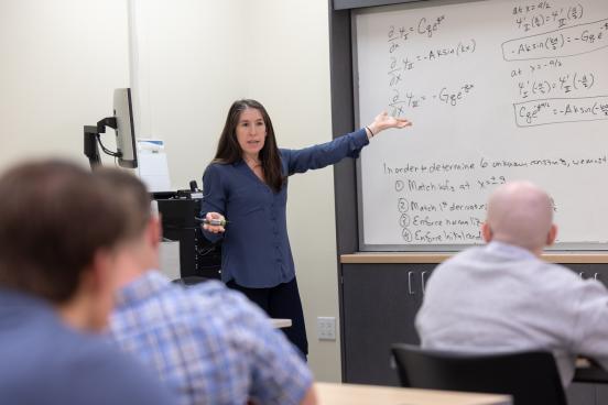 Professor at white board with writing on the board and students sitting at desks looking towards board