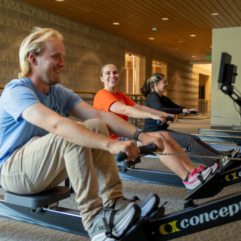 Three students use rowing machines in a campus recreation center, smiling and exercising together indoors.