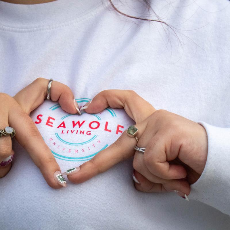 Close-up of a student wearing a “Seawolf Living University” sweatshirt, forming a heart shape with their hands over the logo.