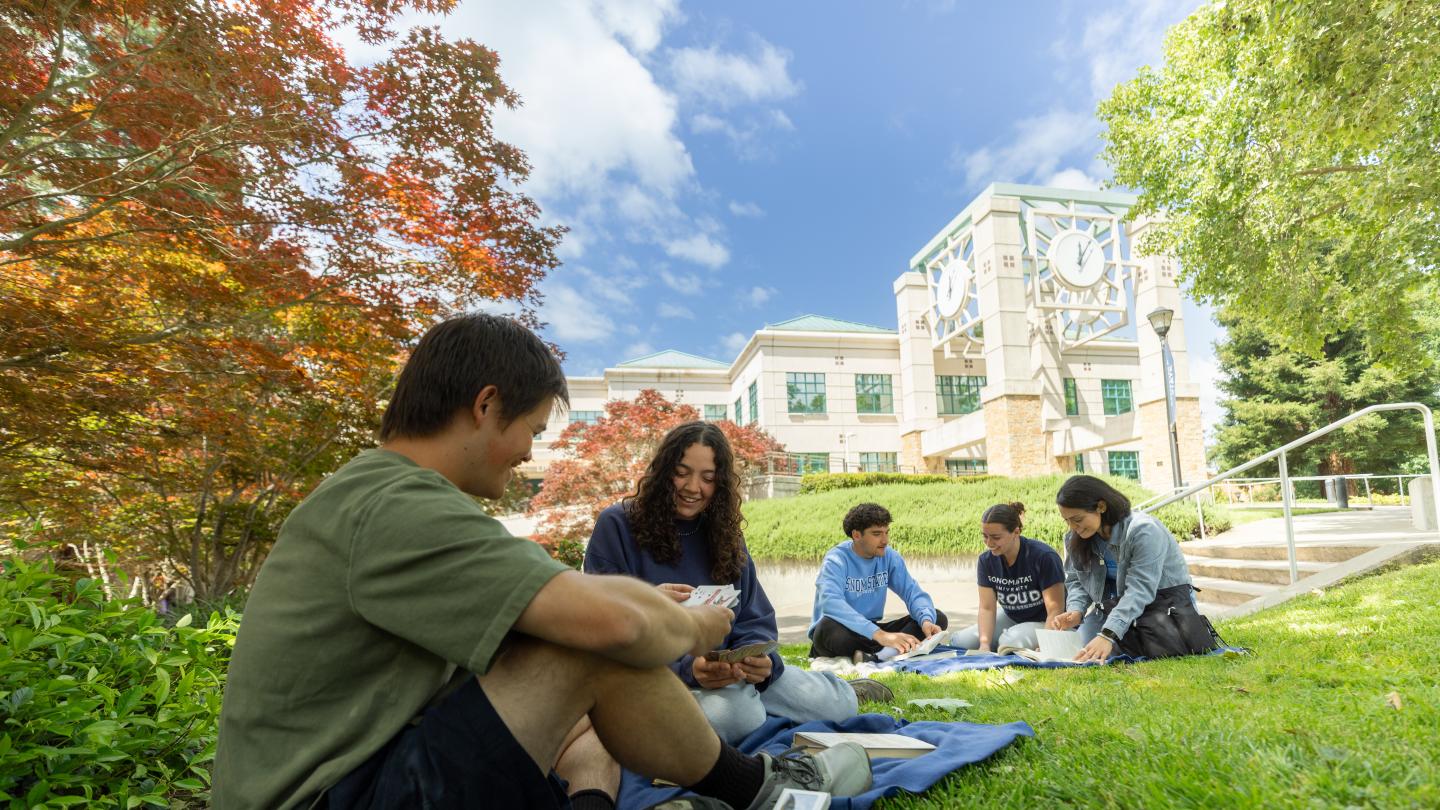 Four students sitting on the grass with the library building in the background