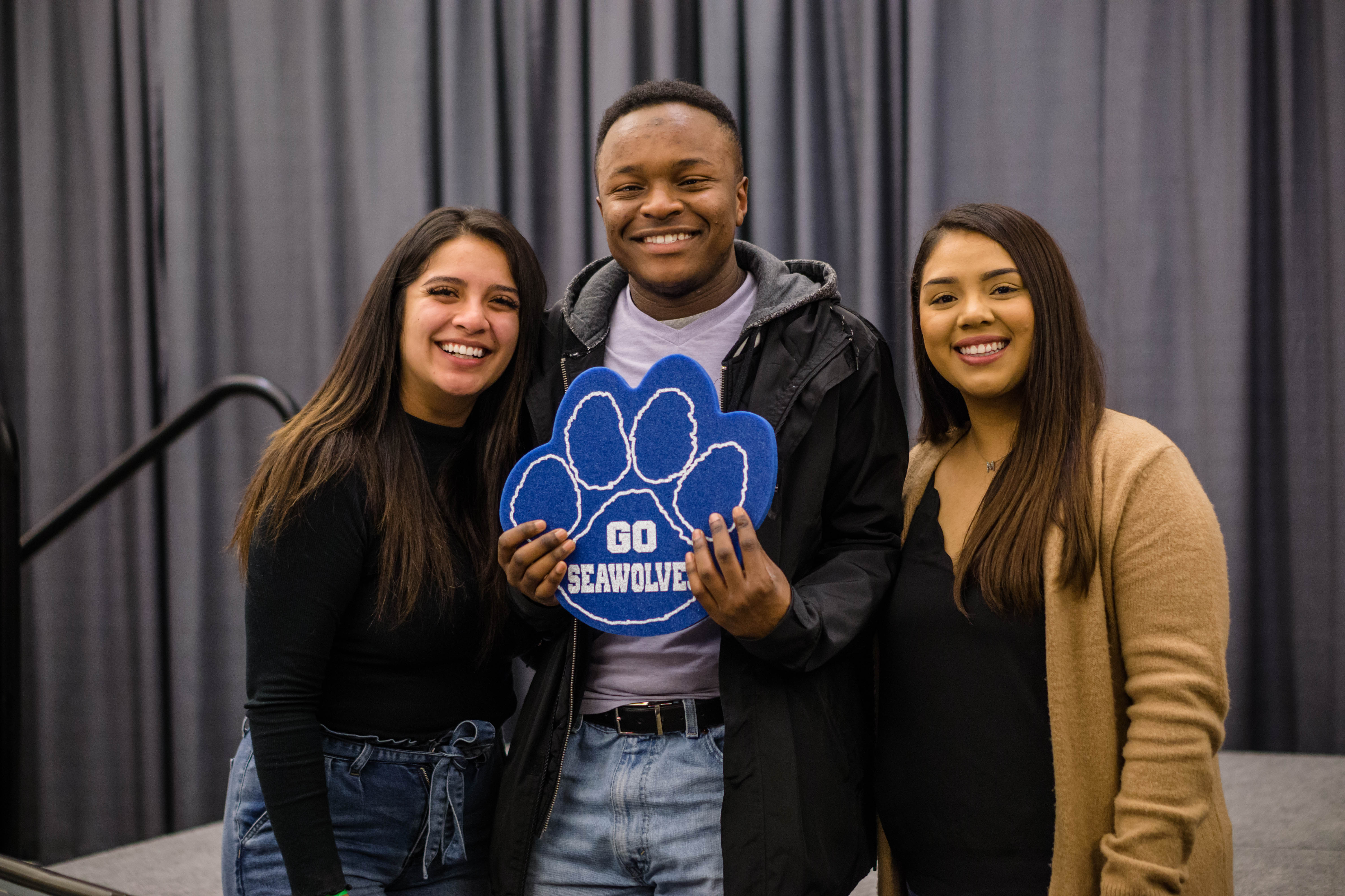 3 students smile at the camera. The man in the middle holds a blue foam Seawolf paw that reads "Go Seawolfs"