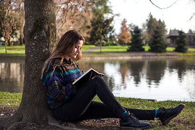 Student sitting and reading a book while leaning on a tree on campus