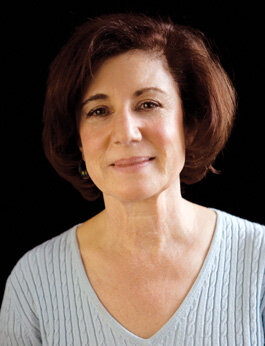 A portrait of Noelle Oxenhandler smiling while wearing a knit sweater in front of a black background