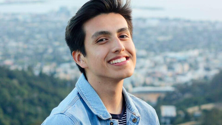 A portrait of Isaias Hernandez smiling in a denim shirt in front of a city skyline
