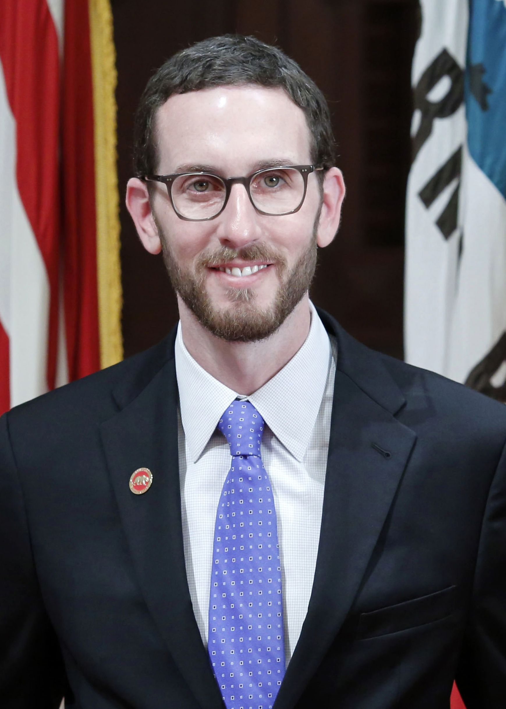 A portrait of State Senator Scott Wiener. smiling while wearing a suit and eyeglasses with flags in the background