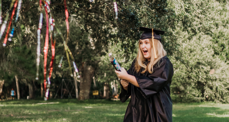 A graduate with a surprised expression popping a confetti party popper while wearing a graduation cap and gown 