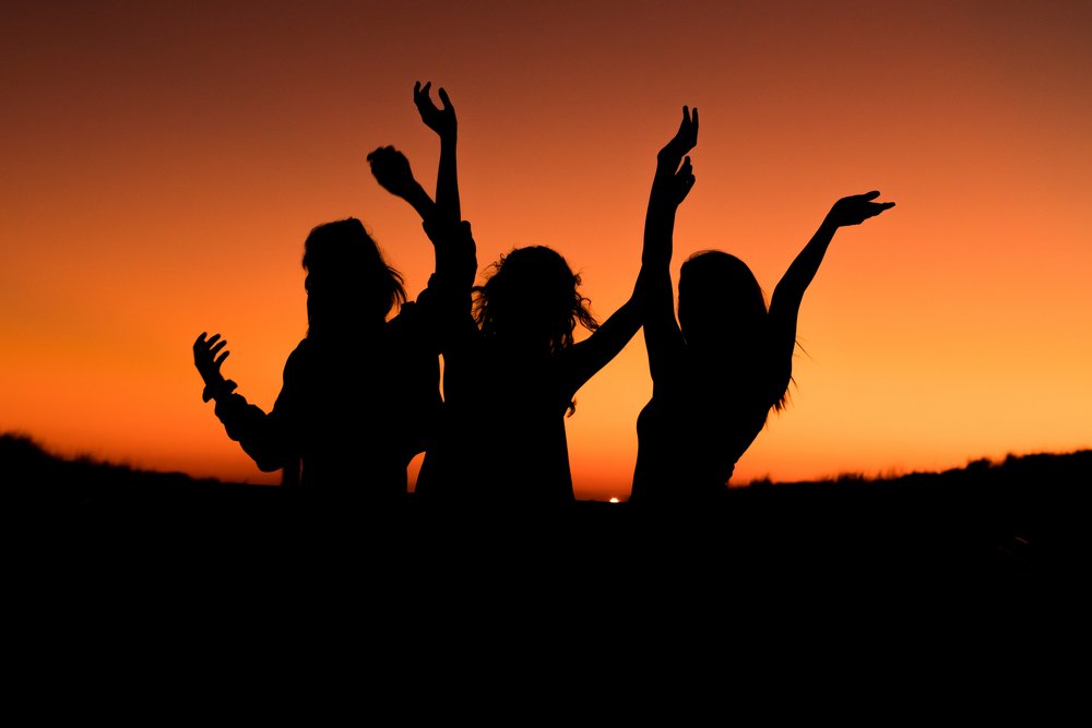 the silhouettes of three people dancing with an orange sunset behind them