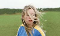 A person with long blond hair blowing in the wind and a blue and yellow shirt standing in a green field