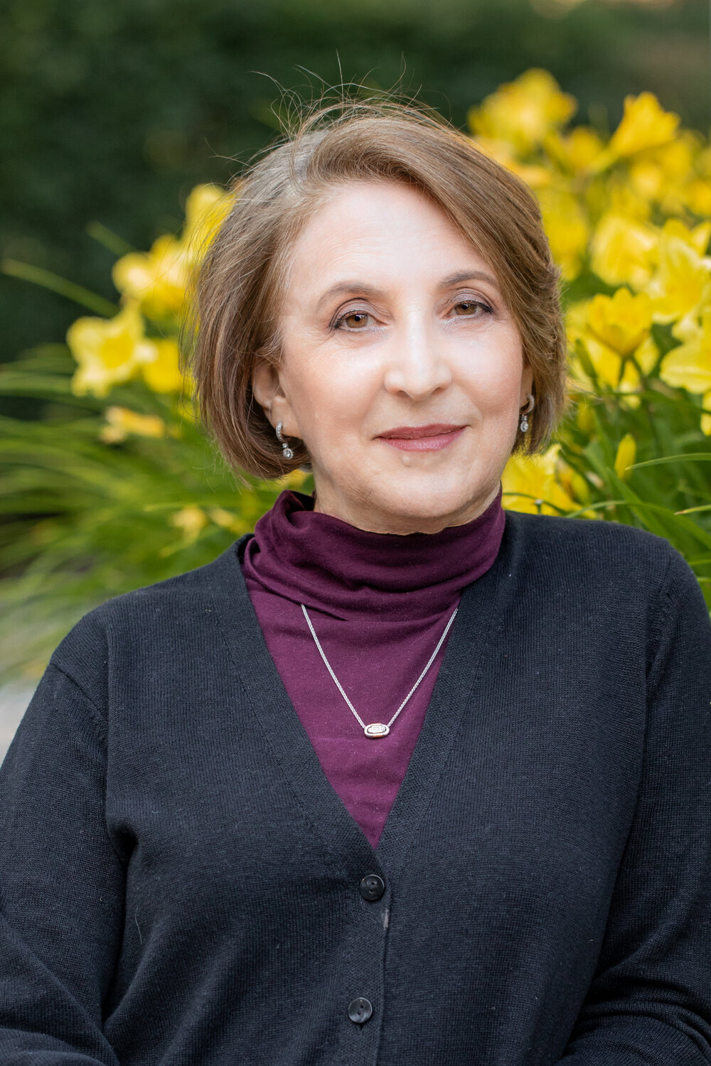 Portrait of Bernice Lerner smiling while wearing a maroon turtle neck, grey cardigan, and gold necklace in front of blurred yellow flowers