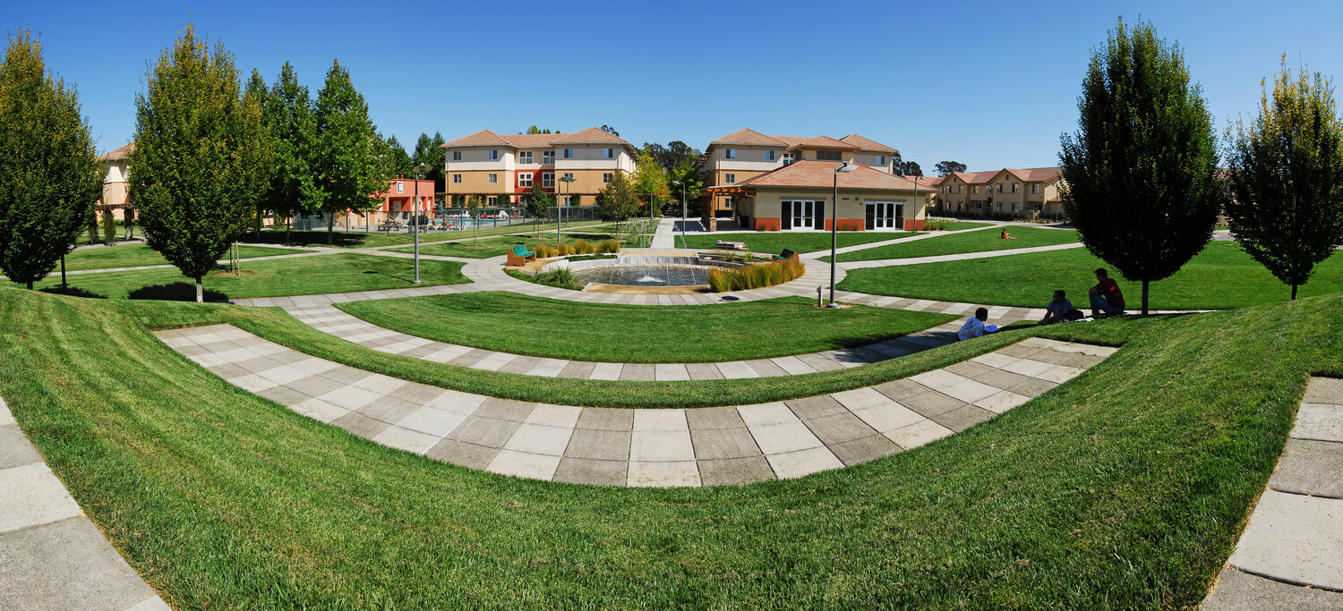 A wide-angle view of the outdoors area of on-campus housing including grass areas, trees, walking paths, and a fountain
