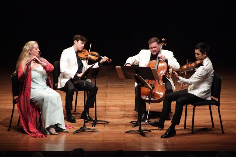 A group of 4 people sitting while playing wind and string instruments on a well-lite stage 