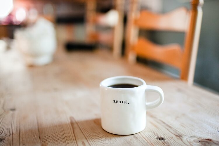 A white ceramic coffee cup with the word "Begin" on it in black letters sitting on a wooden table