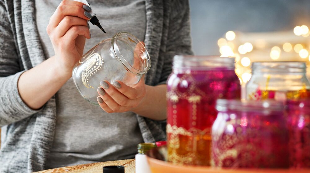 A person in grey clothes writing/drawing in pen on a glass jar with maroon-colored jars in the foreground