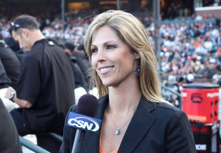 Woman with long brunette hair wearing black blazer speaks into a CSN microphone with large crows of people in stadium in the background