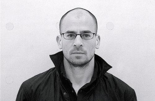 A black and white portrait of Andrew Zawacki wearing glasses and a black collared jacket in front of a white background