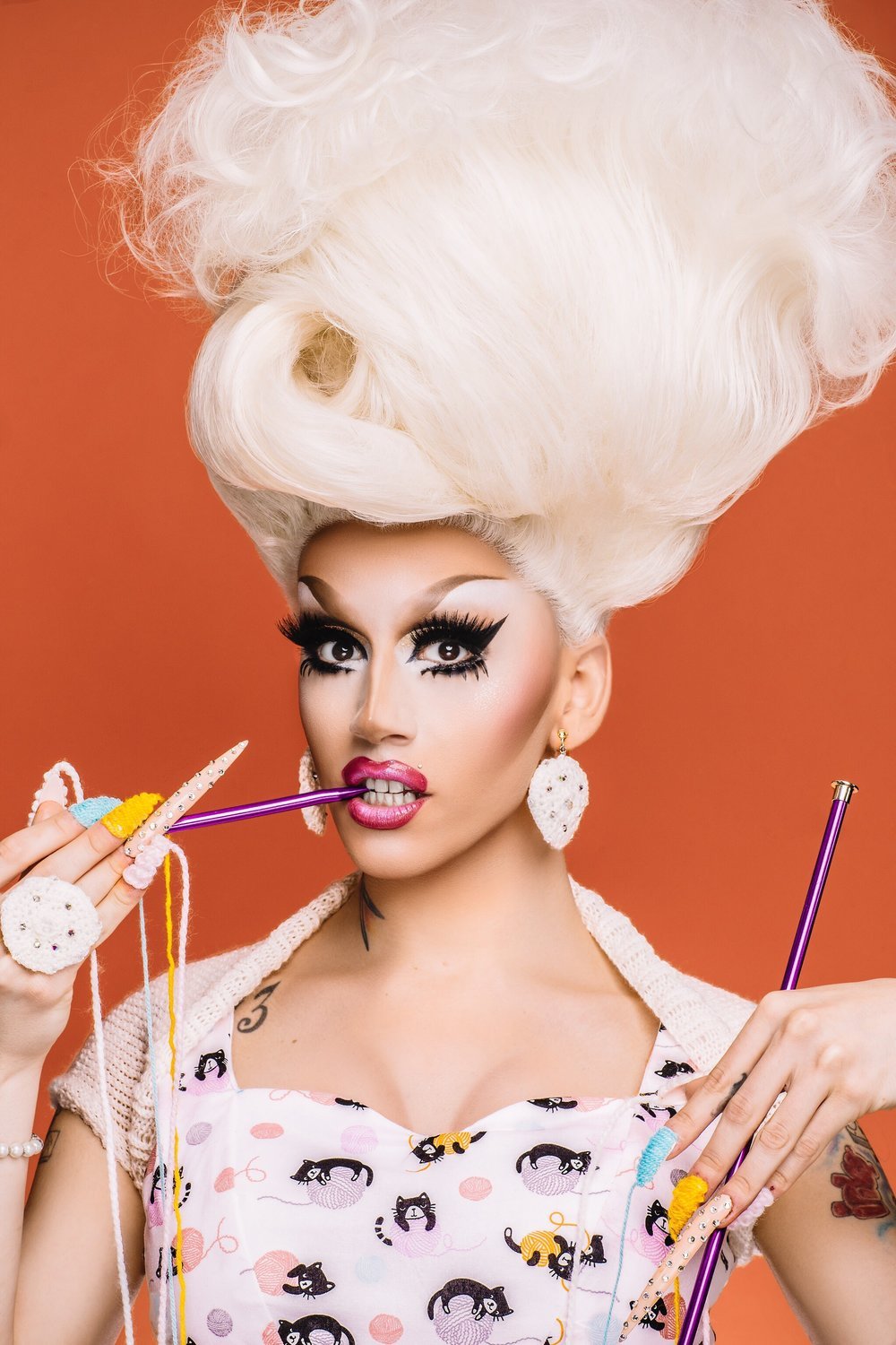 a drag queen biting on a purple metallic knitting needle