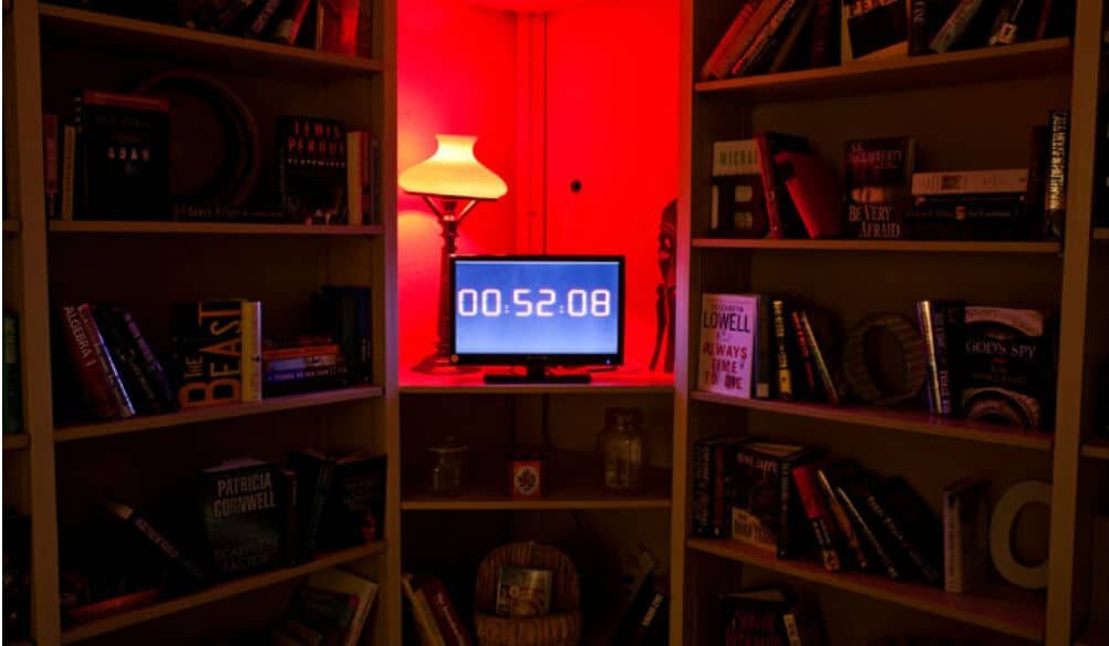 A darkly lit room with filled bookshelves, a red lamp, and a computer screen with the digits '00:52:08' displayed on it