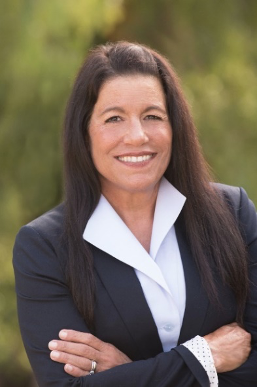 Portrait of Dr. Camille Filardo-Kraft smiling while wearing a black blazer above a white collared shirt in front of a blurred outdoor background