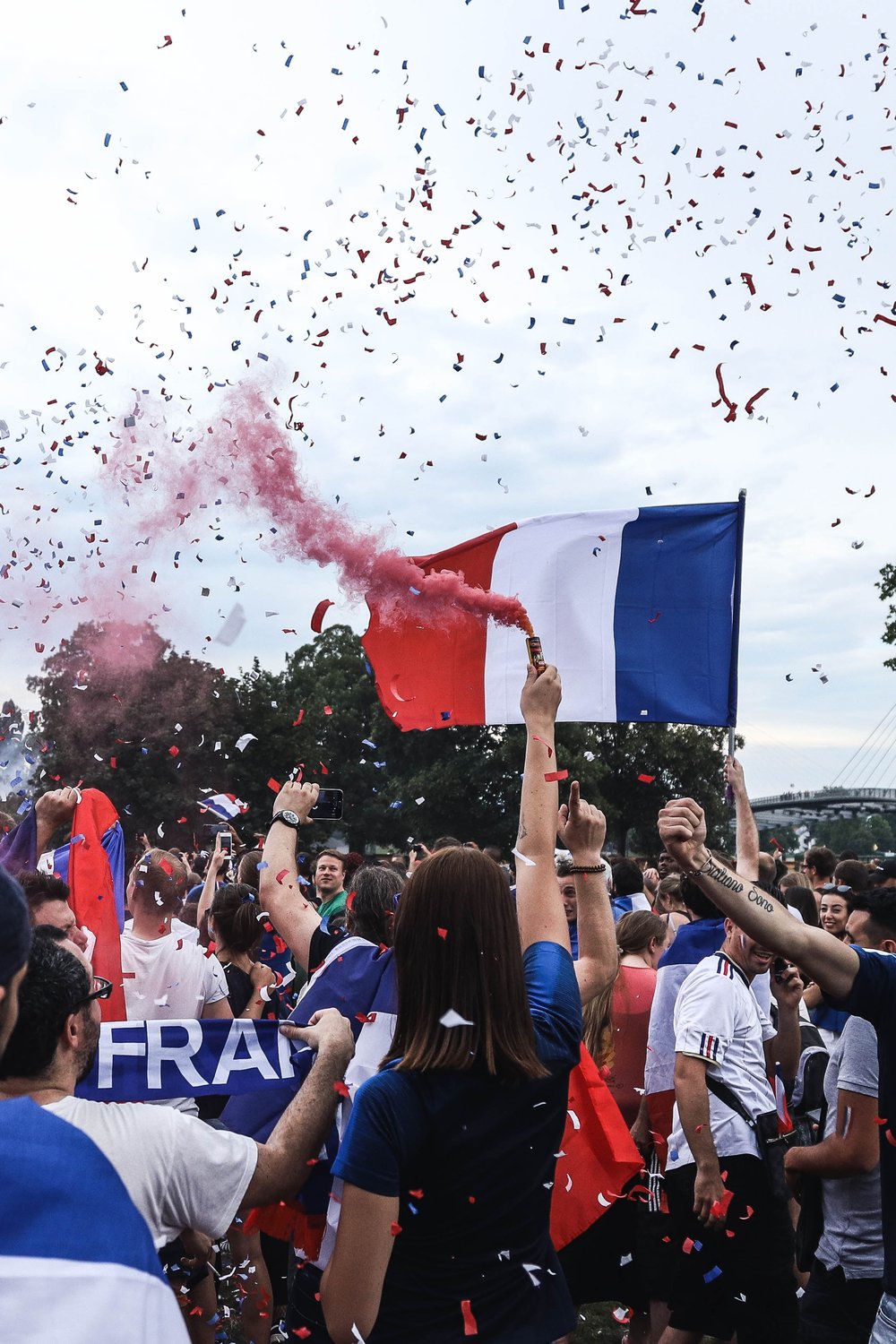 a person holding a red smoke bomb in a crowd of people with another person holding the French flag and confetti falling from the sky