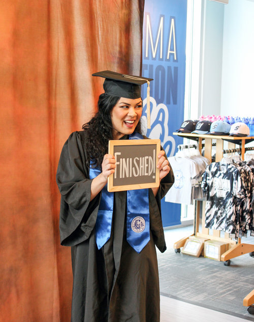 A graduate with long black hair in Sonoma State University regalia smiling and holding a chalkboard with the word "Finished!" on it