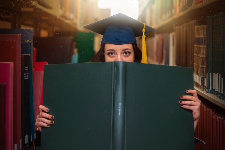 Person inside a library holding a large green diploma in front of their face while wearing a blue graduate cap with a yellow tassel 