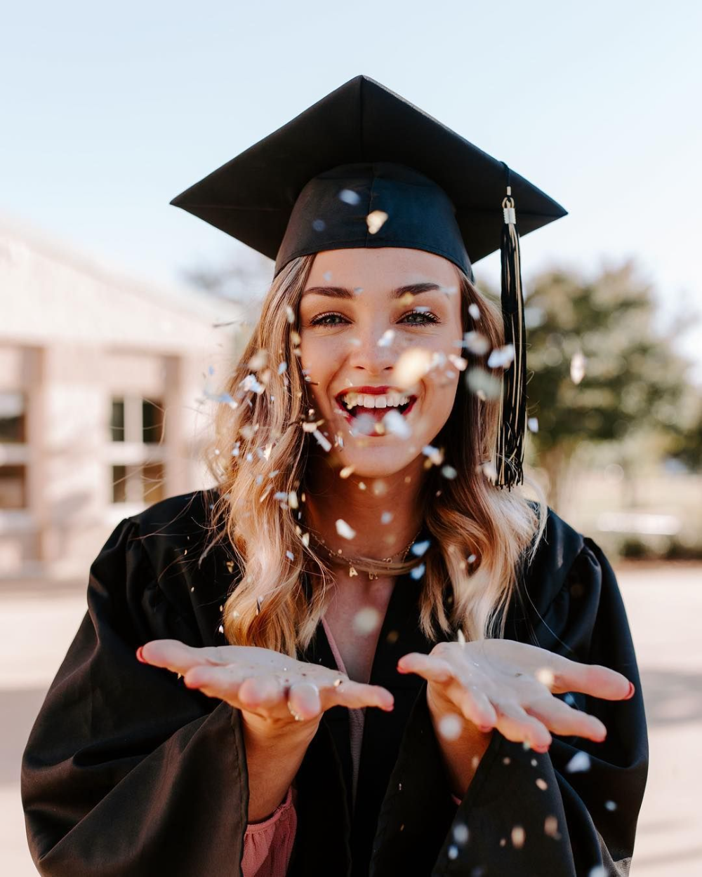 A graduate wearing a black cap and gown smiling and throwing confetti 