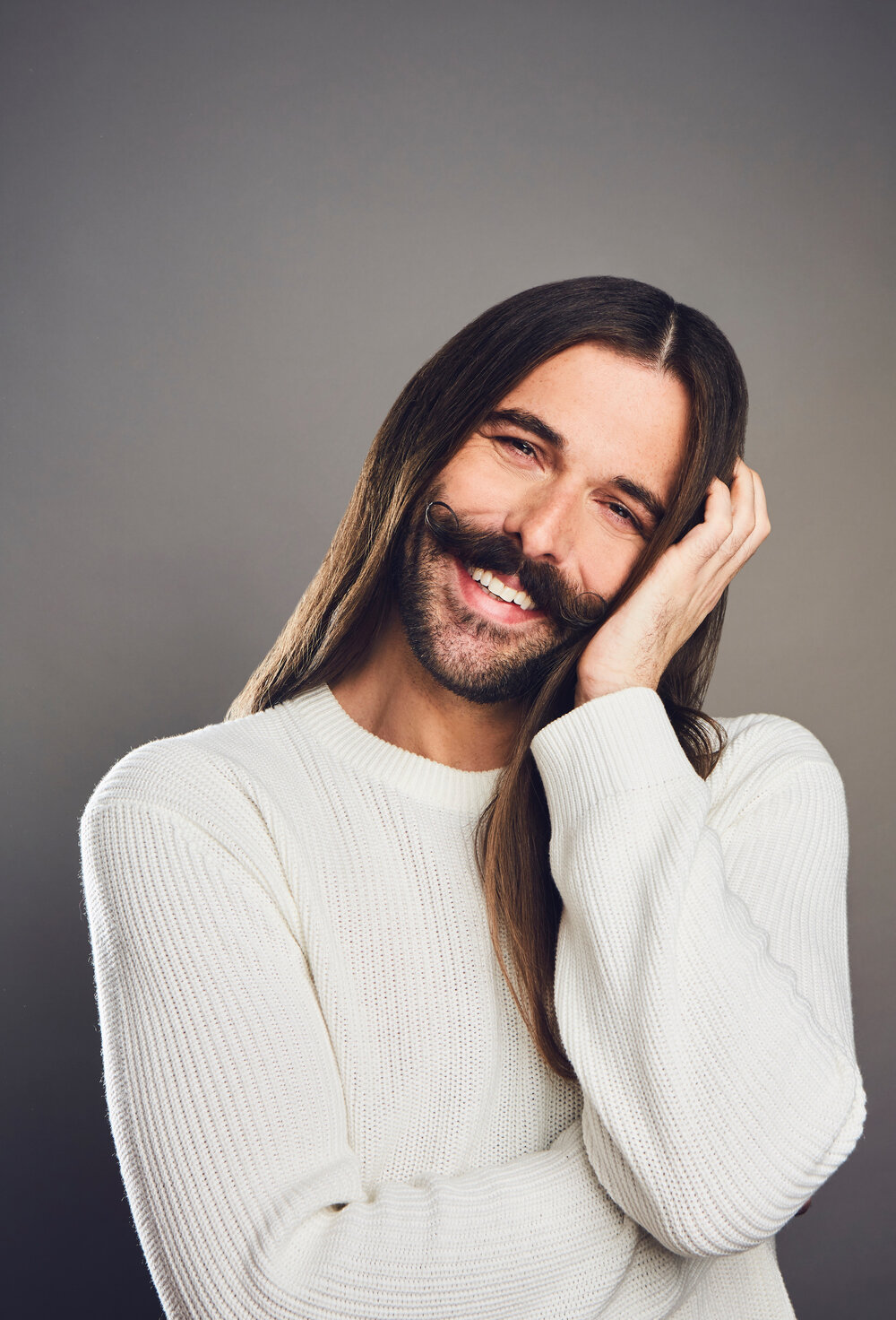 Portrait of Jonathan Van Ness smiling while wearing white sweater in front of a faded grey background