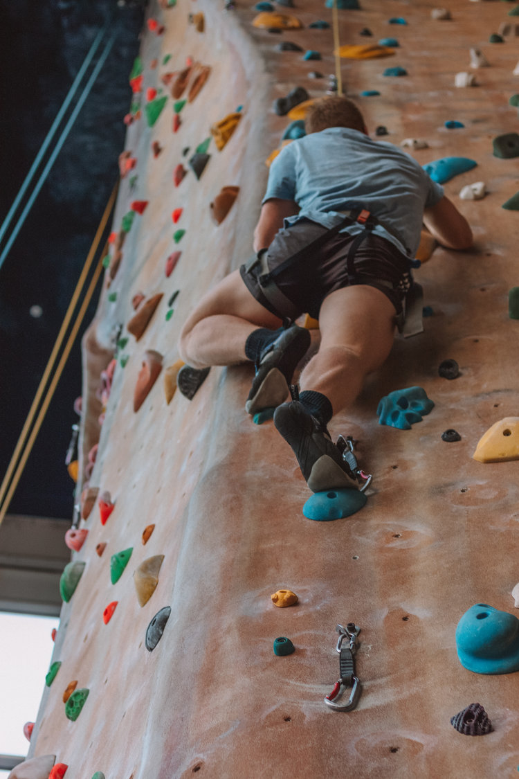 Person climbing indoor rock wall in blue t-shirt and grey shorts