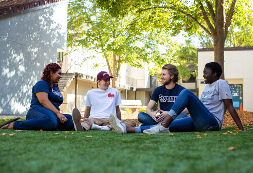 A group of 4 students smiling and sitting on a lawn with buildings in the background