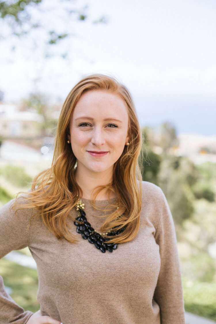 Headshot of Kelly Sullenburger wearing a brown blouse, black necklace, and smiling in front of a blurred natural background