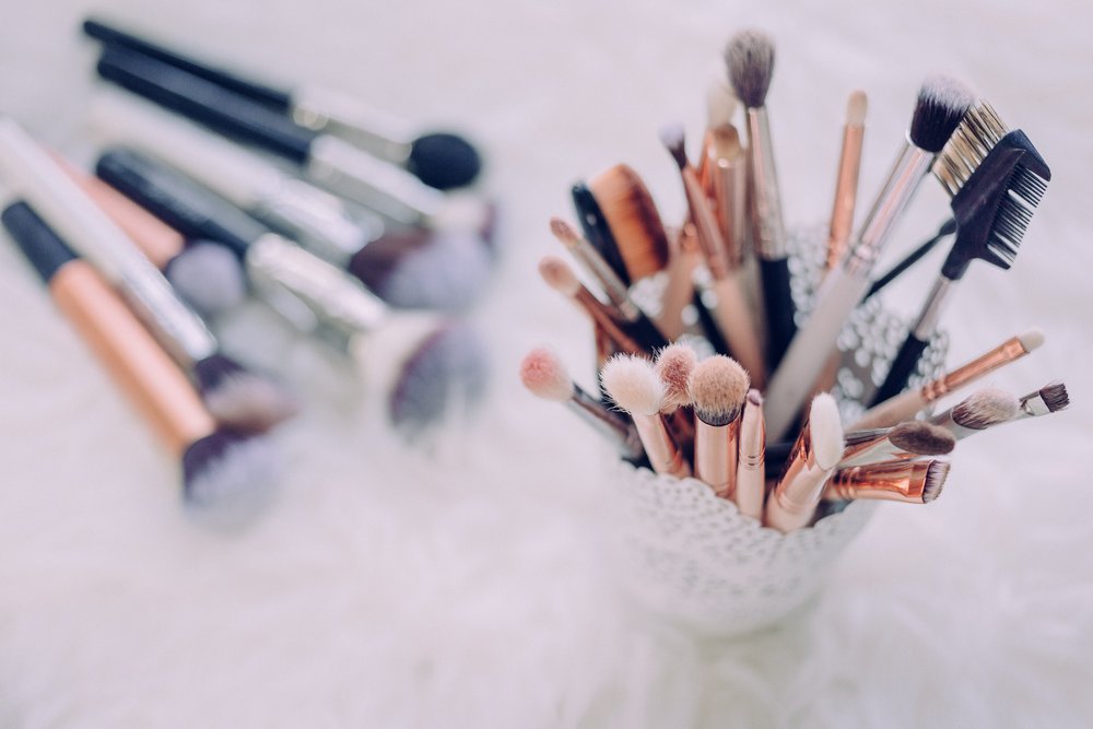 a variety of makeup brushes lying on a flat surface next to a variety of makeup brushes in a cup