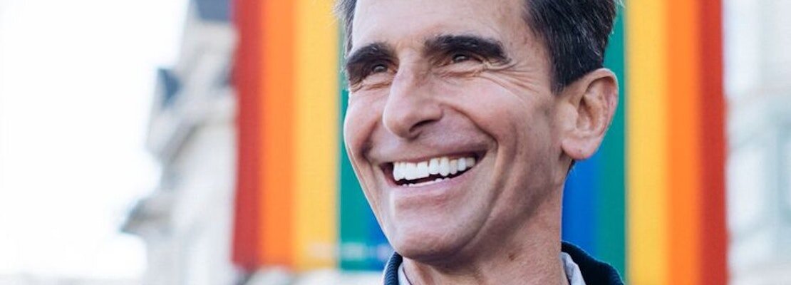 Portrait of former California state senator Mark Leno smiling in front of a rainbow LGBTQ pride flag