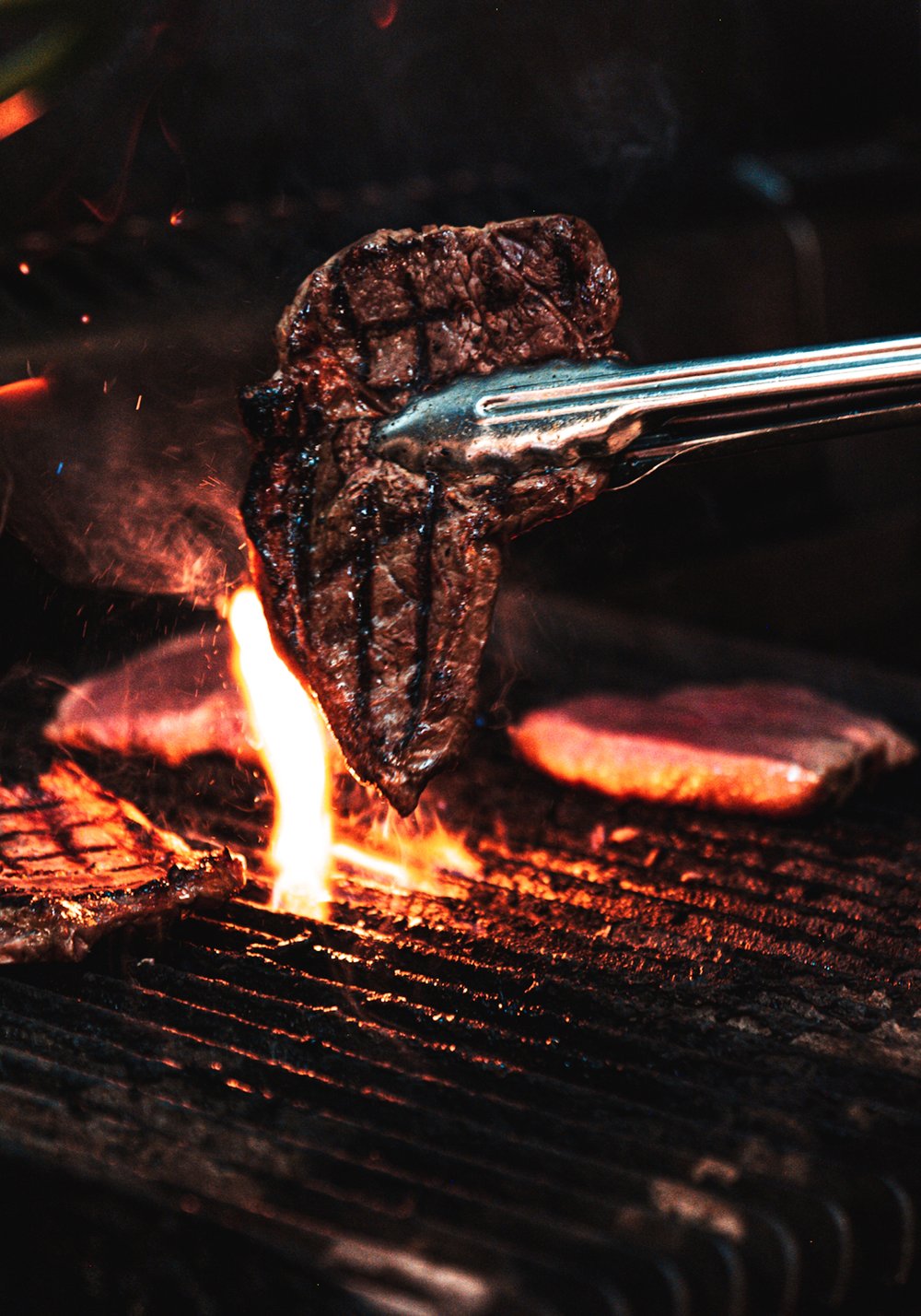 A pair of tongs holding a slab of meat above the flame of a barbecue 