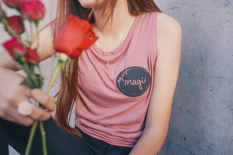 A person with long red hair wearing a pink shirt with the words "Magic" on it holding a small bouquet of colorful flowers