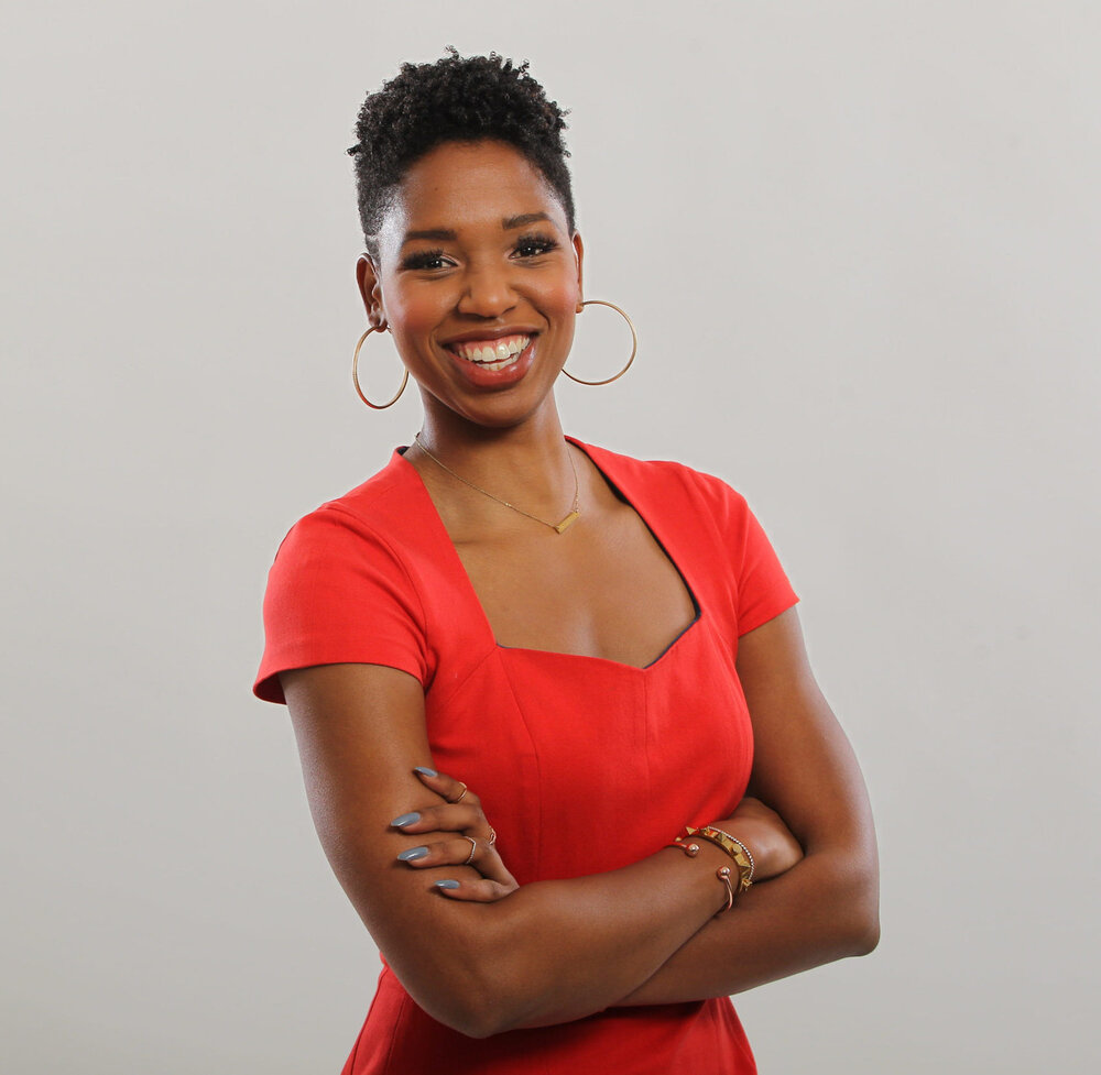 Portrait of Monica McNutt wearing a coral dress and gold jewelry while smiling in front of an off-white background