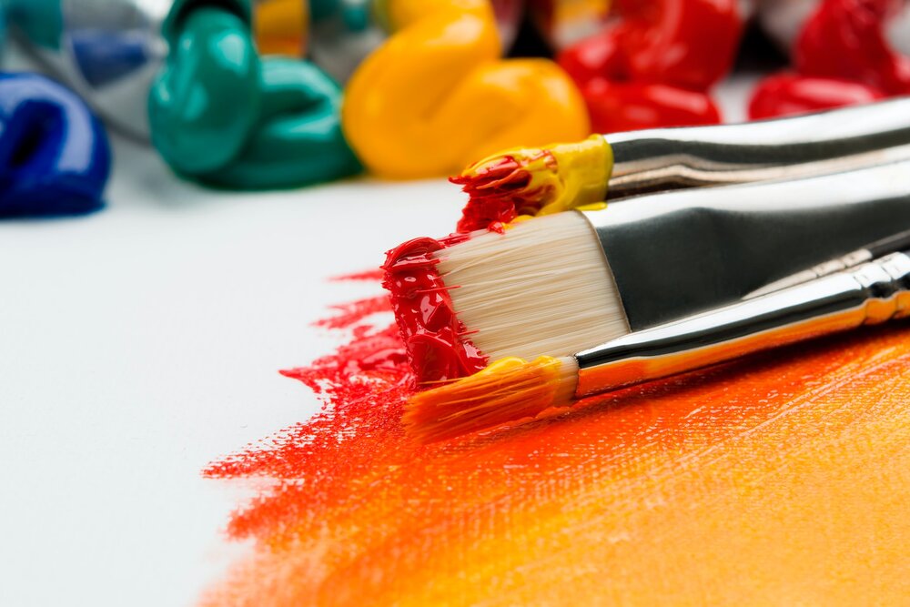 Paint brushed covered in red, orange, and yellow paint with squeezed multi-colored paint tubes in the background.