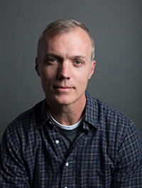 Portrait of Patrick Madden smiling while wearing a dark blue plaid button-up shirt in front of a grey background