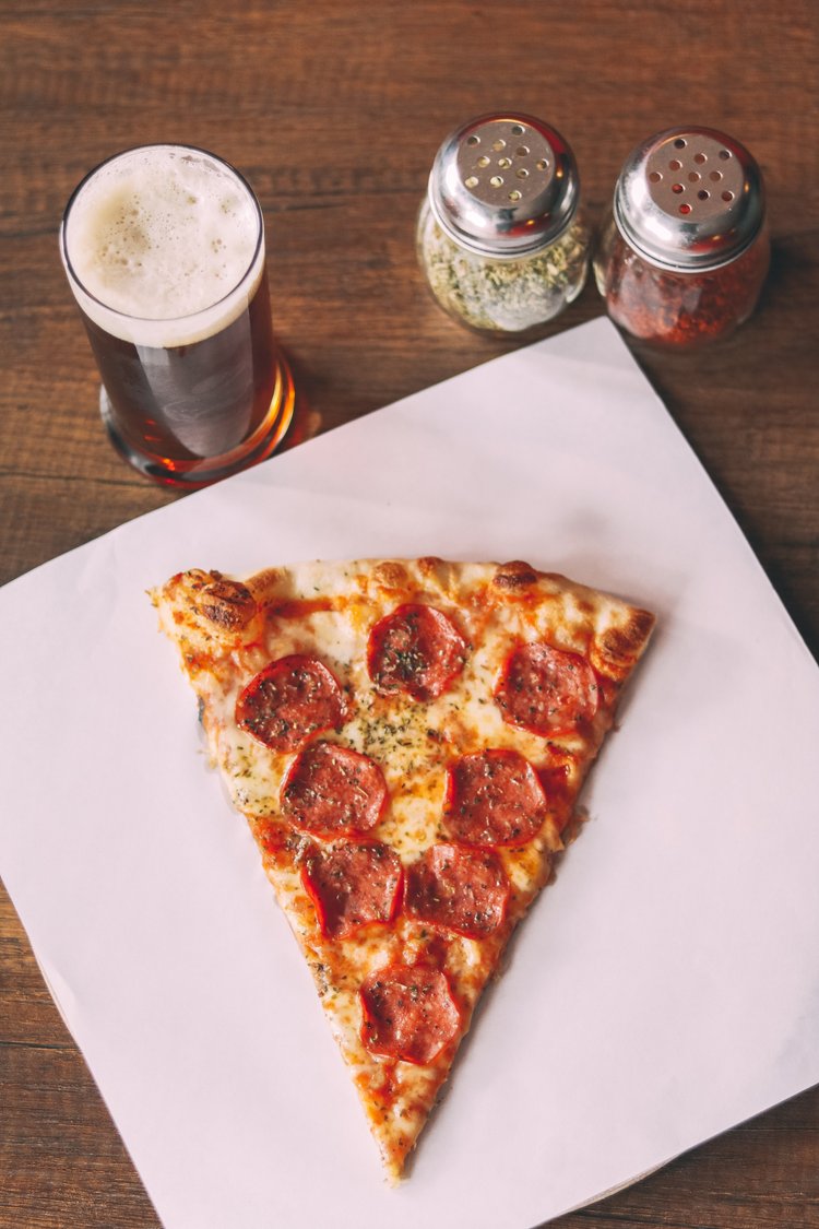 A slice of pepperoni pizza, a glass of beer, and two condiment shakers 
