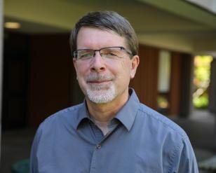 Headshot of Professor John P. Sullins smiling with a blue collared shirt on and frameless glasses.