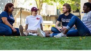 Four students smiling, talking, and sitting in the grass outdoors 