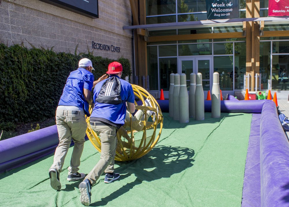 Two people rolling a giant ball towards giant bowling pins in front of the Campus Rec Center