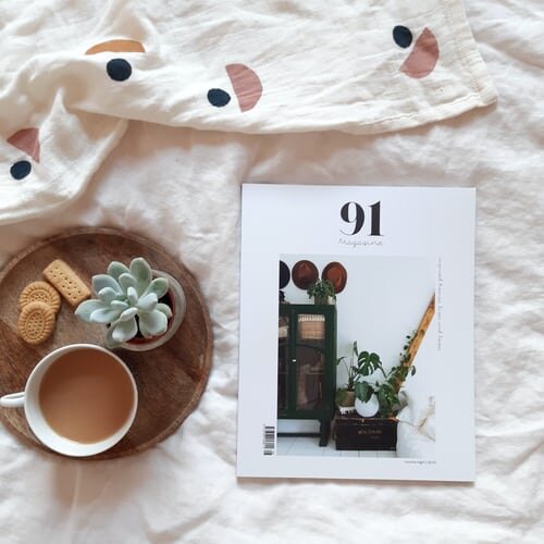 A wooden tray holding a mug of coffee, a small potted succulent, and some cookies lies next to a graphic magazine on an off-white patterned textile