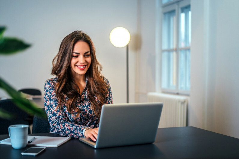 Someone smiling while sitting at an indoor table with a laptop in front of them and a mug, cellphone, and paperwork to their side