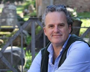 Portrait of Stephen Bittner wearing a light blue collared shirt with a dark green vest while smiling and sitting in an outdoor setting