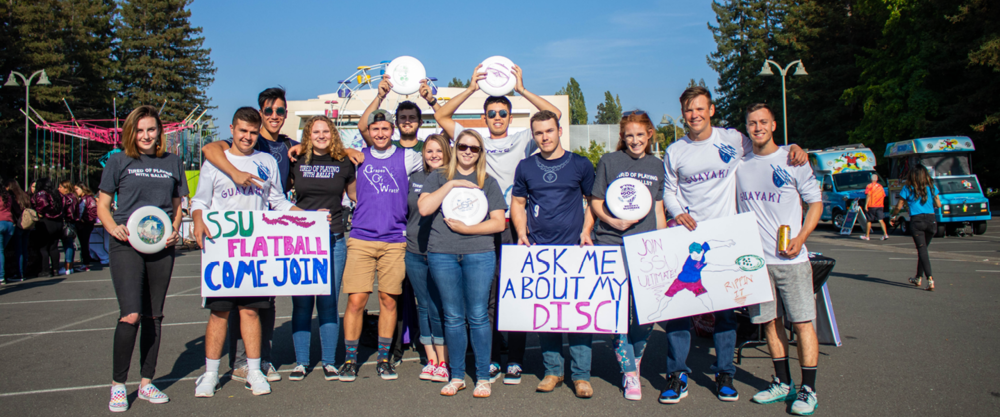 A large group of students posing together outdoors holding colorful handmade signs