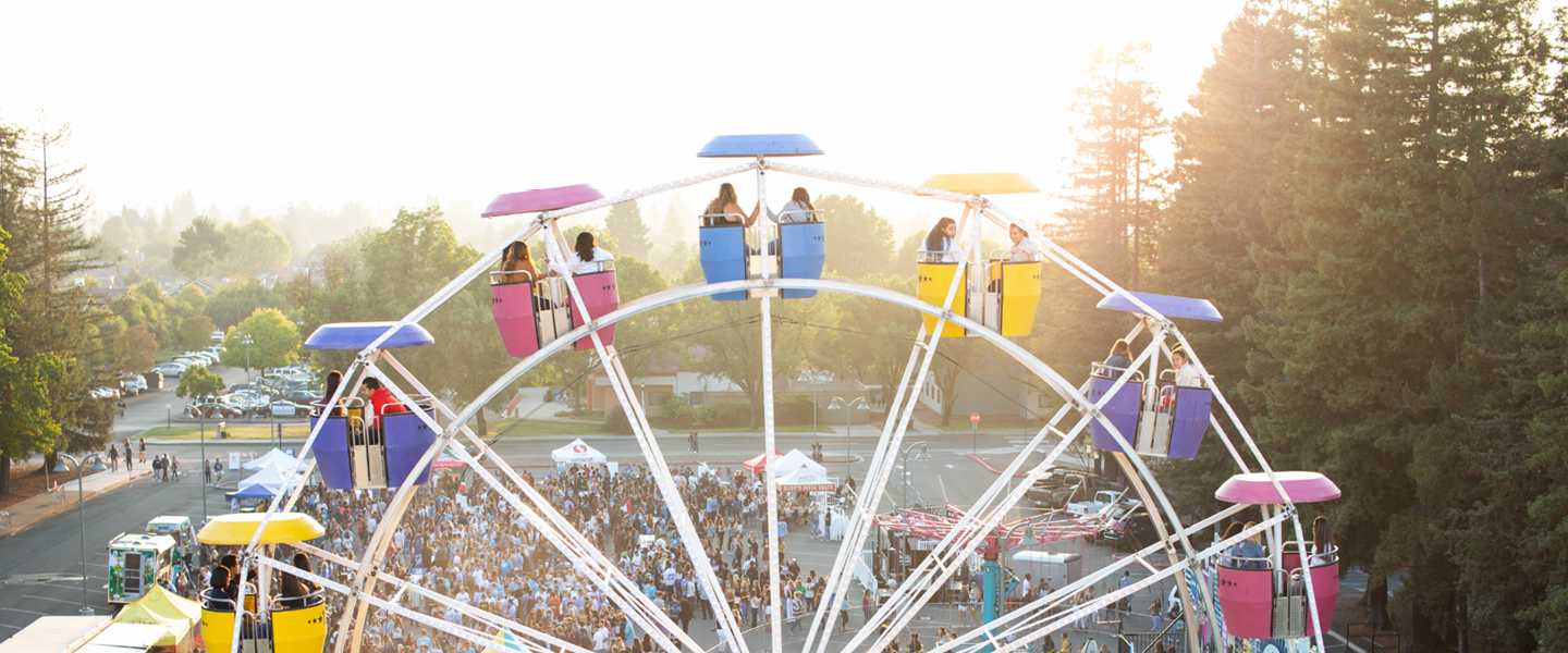 People on a multi-colored ferris wheel with a crowd, trees, and the sunset in the background 