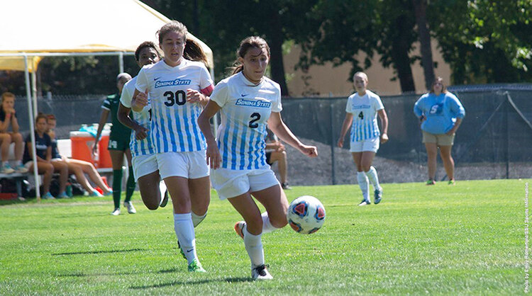 Two people on the women's soccer team kicking a soccer ball while wearing soccer uniforms