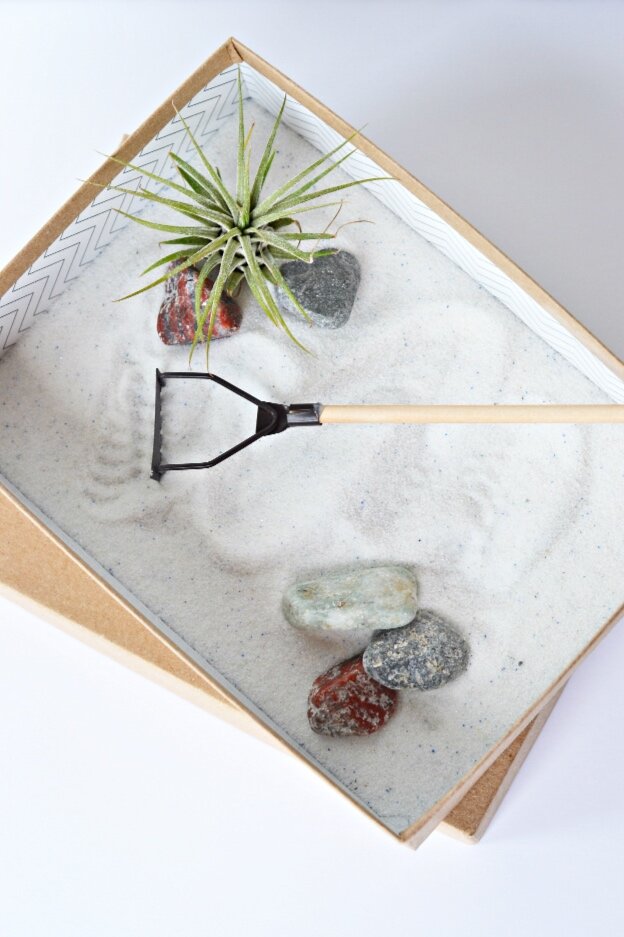 A zen garden including a gold box, an air plant, multi-colored rocks, white sand, and a small wooden rake